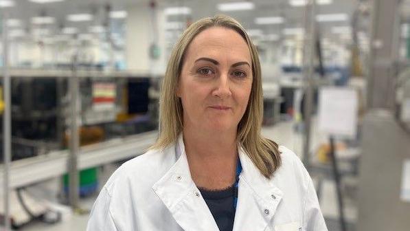 A woman wearing a white lab coat in front of a backdrop of a laboratory