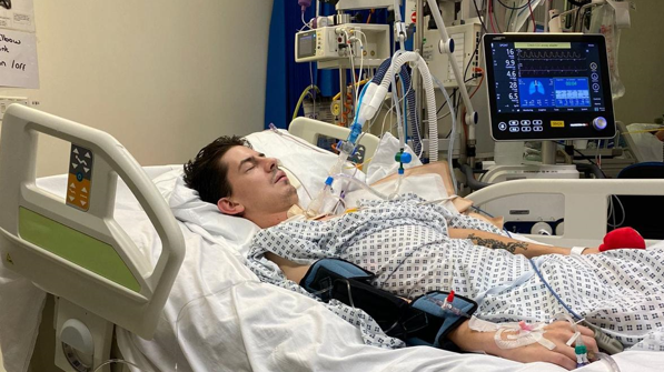 A man lies in a hospital bed, attached to monitors and tubes, with a screen showing information about his condition