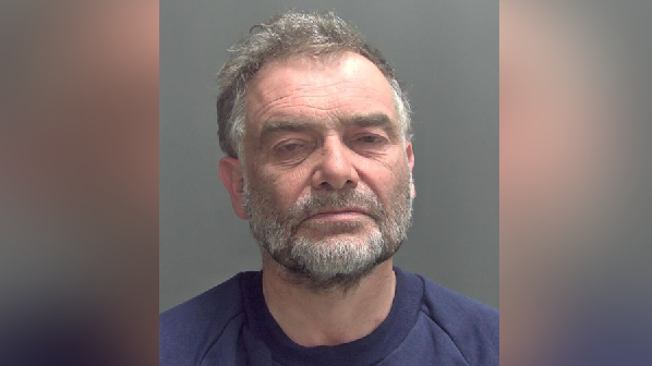 Image shows mugshot of man in purple jumper, he has a grey beard