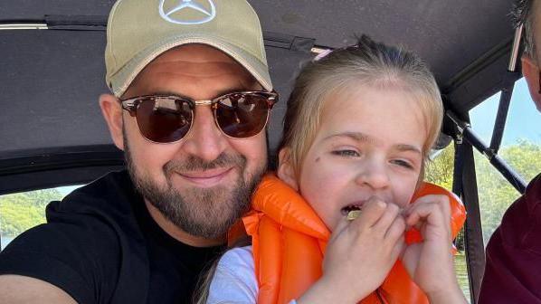 Mr Shreeve, in a beige hat, holds on to his daughter India, who is eating and wearing a life jacket 