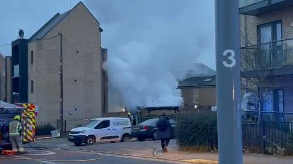 Smoke billowing near blocks of three storey flats. A fire engine cars and fire crews can be seen. 