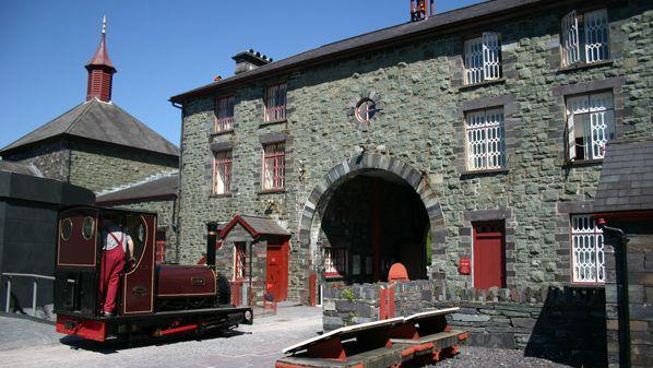 View of the old buildings in the National Slate Museum with another view of that dark red steam locomotive alongside the main entrance arch.