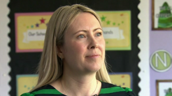Ms Davidson has long blonde hair and is wearing makeup and a green and black t-shirt. She is standing in a classroom and behind her is a black notice board with colourful pages on it.