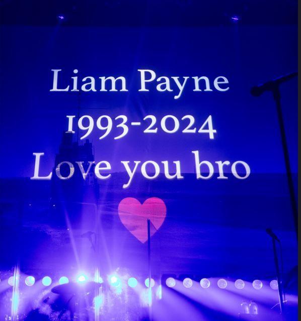 A blue screen with the words Liam Payne, 1993-2024, Love you bro, written in white. A pink heart is underneath and a reflection of stage lights on the surface.