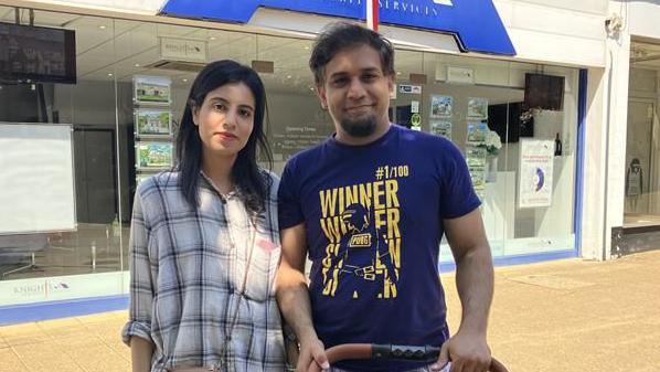 A man and a woman stood outside an estate agents shop with a grey front and blue sign. They are stood in the shade, but the rest of the street is under a bright sun.