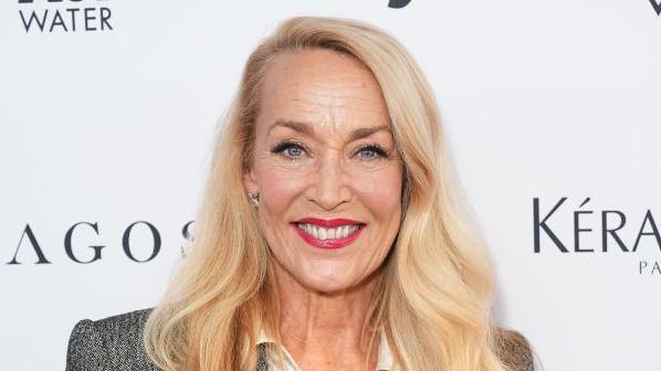 A picture of Jerry Hall, who has shoulder length blonde hair, and is wearing a white shirt and grey jacket.