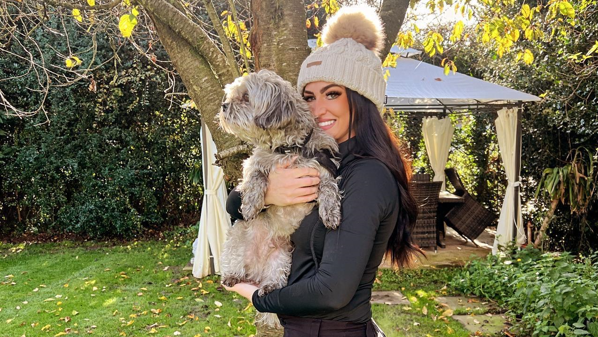 Anna is in her garden holding a dog. It is sunny and she has a bobble hat on and is dressed in all black.