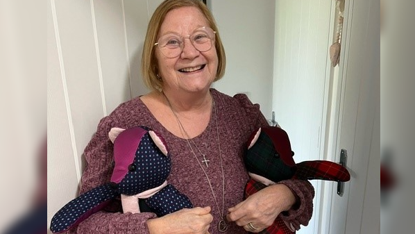 Mrs Taylor wearing a pink jumper, she has medium length blonde hair and glasses. She is smiling. She is holding her two teddy bears. Which are multicolour 