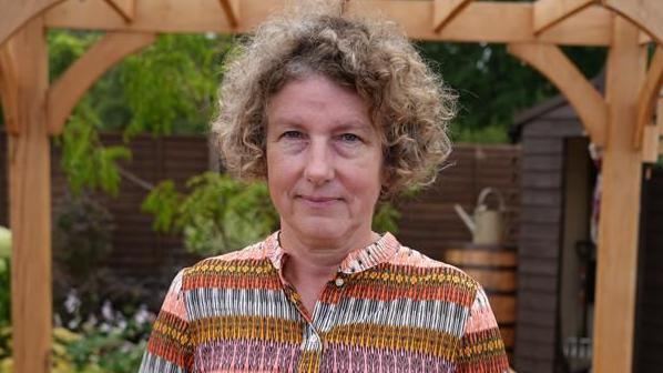Amanda Thomas. A woman who is standing in front of a garden and under a pergola. She is looking directly at the camera and smiling and is wearing a yellow, pink, purple and orange patterned dress. 
