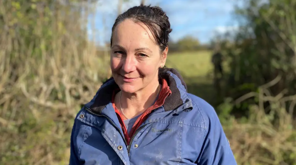 Catherine Withers smiles into the camera, wearing a blue jacket and a field is seen in the background. 