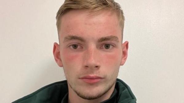 A headshot of Jamie Kadolski looking directly at the camera. He is wearing a green fleece with the collar up. He has short blonde hair. 