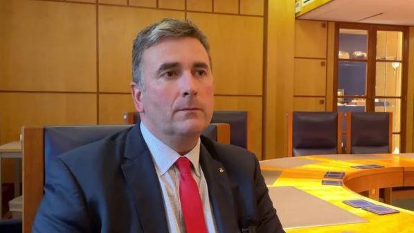 Tim Johnson, who has short brown hair and is wearing a blue suit jacket over a light shirt with a red tie. He is sitting at a large table in an office.