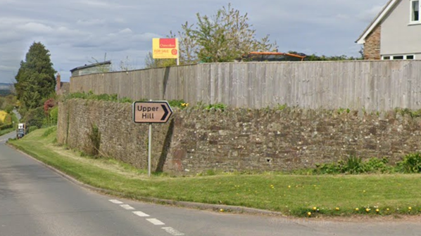 A road junction with an arrow shaped sign that reads "Upper Hill". A stone wall follows  around the corner of the junction. The sky in the background is grey. 