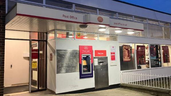 The entrance to the post office which features an ATM and a post box on the right. Several posters hang in the window as well as a currency conversion board.