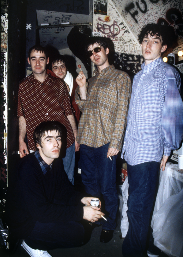 Colour photograph of Oasis posing for the camera. All of them are stood up with the exception of Liam Gallagher who is crouched down.