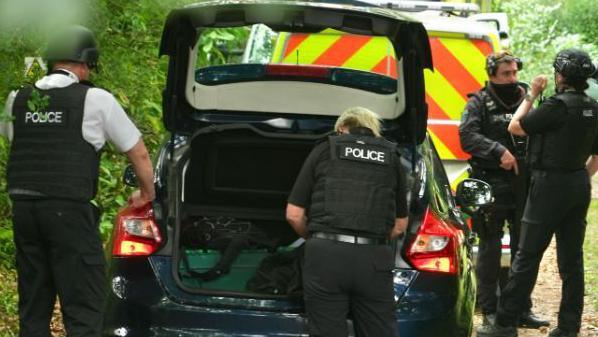 Police officers in stab vests - and one carrying a gun - unload things out of a car boot