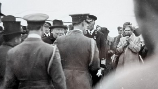 Sir Winston Churchill dressed in dark clothing and wearing his distinctive Homburg hat, meeting senior military officials, all in uniform.