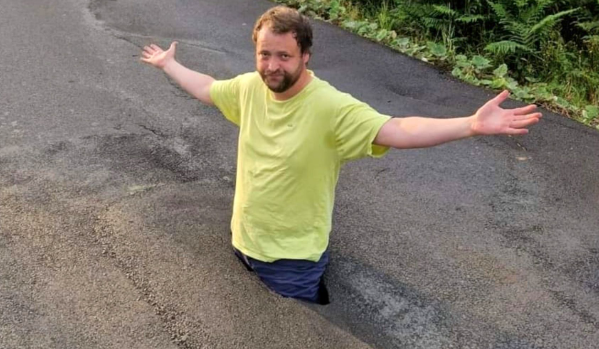 A man stands waist deep in a pothole in the middle of a small road during the daytime. He is wearing a yellow t-shirt, blue jeans and has his arms outstretched.