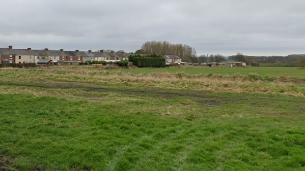 The land for the proposed housing development which is a large, open field with terraced houses visible in the background.
