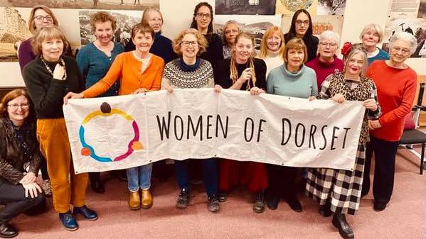 A group of 17 women posing for the camera. The people in the front of the group are holding up a long white banner saying Women of Dorset.