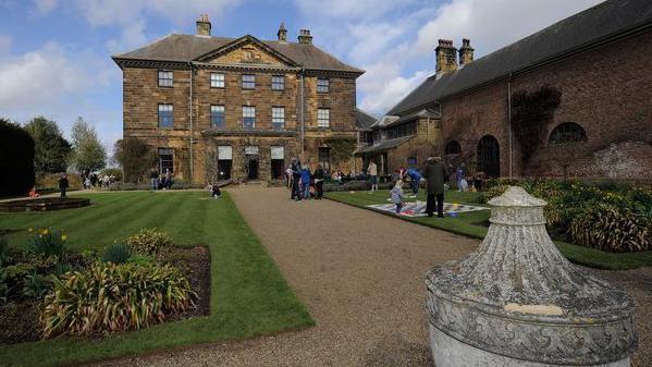 View of Ormesby Hall, a classic Georgian mansion. There are five windows on each of the top two storeys and a door and a portico aligning with the roof above the front entrance. Members of the public are strolling on the  gravelled path leading up to it, which is fringed with a well tended lawn and flower beds on either side. The top of a stone urn can be seen on the lower right of the image.