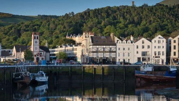 Buildings on a quayside in Ramsey behind boats on the water in the harbour. There are reflections of the buildings on the water and the scenes has a backdrop of hills covered in trees