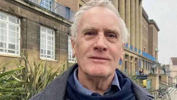 Councillor Mike Sands stood outside Norwich City Hall. He has short grey hair, is cleanly shaven and is wearing a grey jacket over a blue jumper and patterned shirt