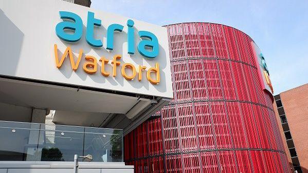 The exterior of a modern shopping centre, with "atria Watford" in blue and orange lettering on a white panel above the glass wall of a shopping centre cafe. There is a circular red wall to the right.