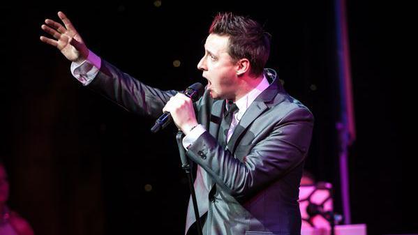 James Huish wears a grey suit, white shirt and black tie. He has gelled up brown hair and his left arm is holding a microphone to his mouth as he sings. His right hand gestures into the air