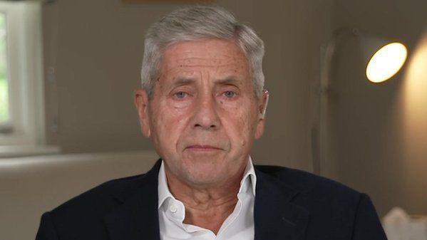 Stuart Rose looks into the camera while sitting down inside a room. He has short grey hair and is wearing a black suit jacket with a white shirt underneath. A switched on lamp can be seen behind him as well as a window.