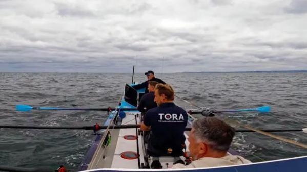 The camera is behind four men in a rowing boat. Blue blades of oars can be seen above a choppy sea.