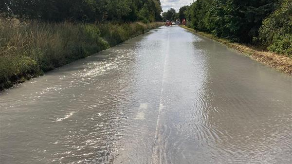 A road pictured from the middle of the carriageway, disappearing into the distance. It is completely underwater and you can barely make out the white lines. It is hedged by verges and trees on both sides and in the distance is a series of vehicles.