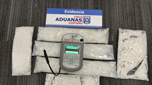 Bags of white powdery substance laid on the floor, with some sort of testing device placed on top of it. Above the photo is a card which says 'Evidencia, Chile Aduanas Customs'.