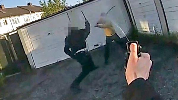 A still from police bodycam footage of a police officer holding up a baton as he confronts a man holding a long curved sword and wearing a yellow hooded top. The arm of the officer who is wearing the camera can be seen spraying pepper spray