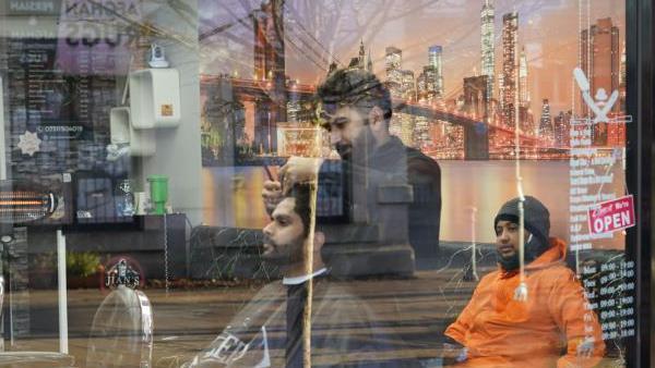 a barber cuts the hair of a customer while another man waits his turn and watches from a sofa