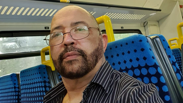 William Waheed Lyall sits on a blue and navy dotty bus seat with yellow handles at the top. The selfie photo shows he is wearing a black and grey and white stripey shirt. He has a beard and thinly rimmed glasses. 