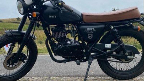 A motorbike with a brown seat, pictured against the backdrop of a country lane and fields.