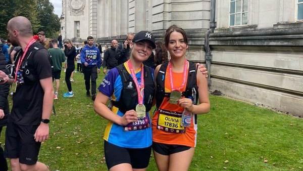 Olivia Browne and her friend Grace Davies stand in front of Cardiff's National Museum. They are holding medals. Olivia is wearing a pink shirt and a black running vest. Grace is wearing a blue shirt and a black running vest. Both have half marathon labels with their name and number on.