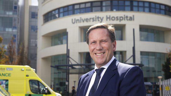 Prof Andy Hardy stood in front of a cream stone hospital building and an ambulance