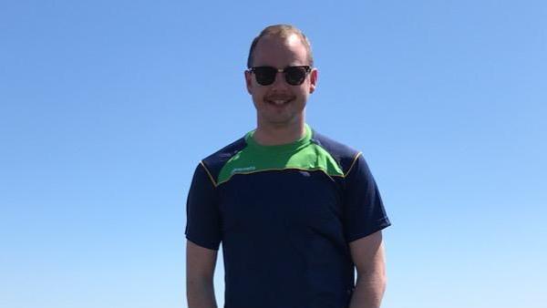 Craig Ballentine smiling at the camera on a sunny day.  He has short, fair hair and a moustache.  He is wearing sunglasses and a navy and green T-shirt.