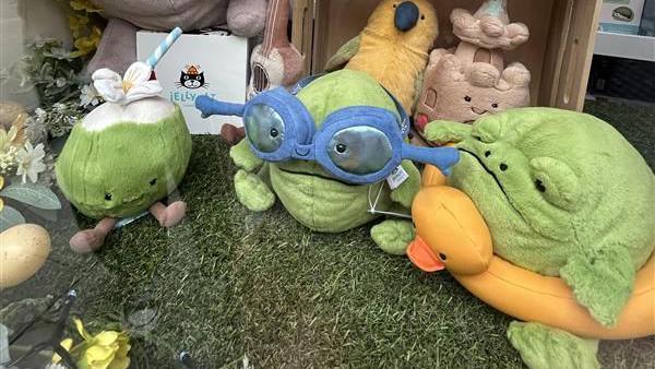 Inside a shop window are Jellycat toys lined up on a grass display. On the left is a green and white drink with a face and brown feet and a straw. In the middle is a frog wearing blue goggles and on the right is another frog with a grumpy expression wearing a yellow duck dinghy. Behind is a sandcastle with a smiley face and a yellow bird next to it.