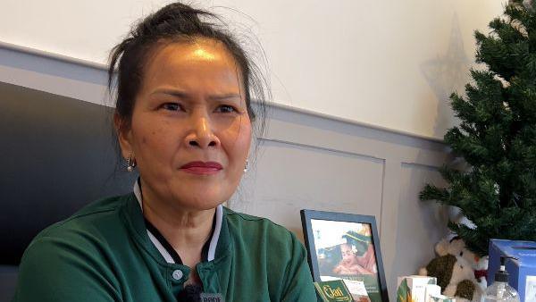 A woman frowns as she sits in a shop in front of a white a grey-panelled wall. A small Christmas tree, framed picture and other items can be seen to her right. She is wearing a green polo shirt and silver earrings. 