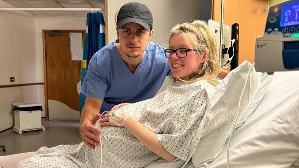 Ace and Shannon during the last moments of pregnancy. Shannon is in a white hospital gown on a hospital bed and Ace is wearing a blue medical gown with a cap on, both looking at the camera with hospital machines behind Shannon