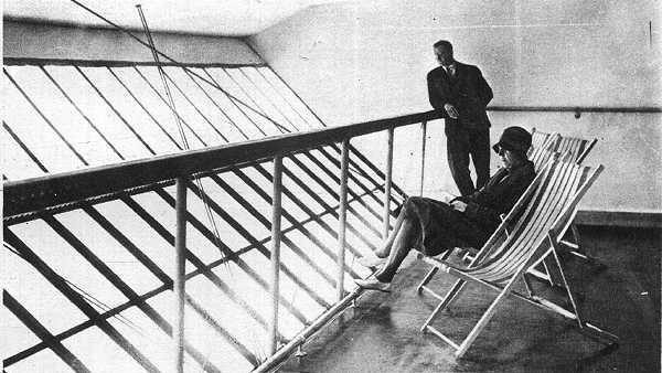 A black and white image of two people behind a barrier on the deck of an airship. On the other side of a barrier are slanted windows. A man is standing up and leaning on the barrier, looking out of the windows. A woman with a hat on is sitting on a deckchair looking out of the windows, with an empty deckchair next to her.