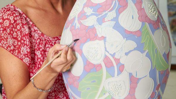 A woman decorates a pottery vase with a flower design on it. We do not see her head but she wears a red dress with white flowers on it and holds a paintbrush with the tip on the vase. Parts of the vase are not painted but some have red, green and blue paint on already.
