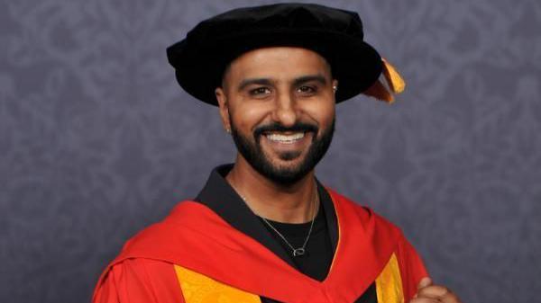 Harpreet Singh Jandu wearing a red and orange gown, holding a graduation scroll