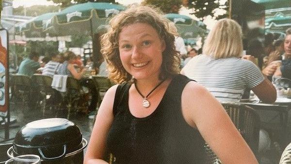 A woman sits at a table in a Parisian outdoor cafe and smiles for the camera. She is surrounded by people seated at other tables. She has curly brown hair and wears a black sleeveless top and a gold pendant around her neck.