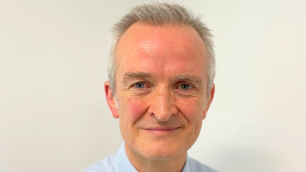 Dr Ewen Cameron is smiling at the camera. He has grey hair and is wearing a light blue shirt and standing in front of an off-white coloured wall.