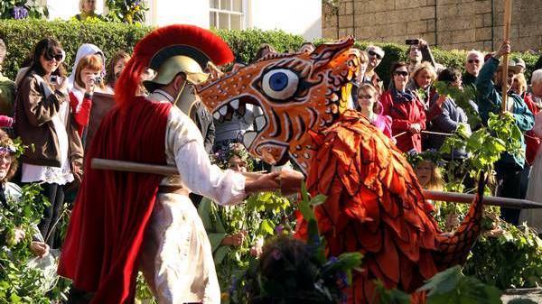 An actor dressed as a Roman with a helmet and cloak is jousting an actor wearing a dragon costume with a paper mache head. The majority of the people in the crowd surrounding them are smiling.