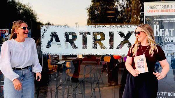 Ruby Tapp (left) and Danielle Jade (right) stood outside of a building. There is writing on the glass window behind them which reads 'ARTIX'. Danielle is holding a flyer with the words 'KIN' on it.